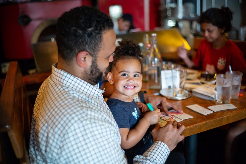Father and young daughter are sitting at a table together in a restaurant. Father is looking down lovingly at daughter. Daughter is looking up at her father with a big smile. 